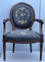 Front view of French style armchair showing round backrest with floral needlepoint upholstery and carved wooden frame and armrests.