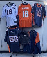 Four Denver Broncos jerseys with tags, one windbreaker, and one backpack displayed hanging and organized to show front and back