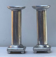 Photo of two tall metal candleholders side by side showing their polished cylindrical columns and square bases with four small feet