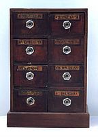 Front view showing the 8 drawers with drug name labels and glass knobs in a dark wooden tabletop chest.