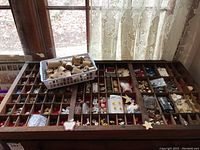Overall view of the large multi-compartment wooden drawer filled with buttons with a white basket of wooden spools placed on top, window and lace curtains in the background.