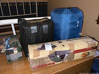 Four camping items on wooden table including tent box, sleeping bag, cot and air pump box