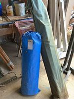 Blue tarp bag and green canvas bag standing upright on a dirt floor with other tools in background