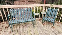Glider bench and chair with matching green metal strap construction set on a wooden deck with fallen leaves around.