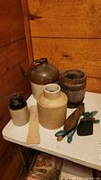 Photo of three vintage stoneware jugs, small wooden barrel, wooden shoe shapers, vintage slide rule, and metal cow bell on a table against a wood wall.