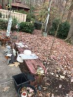 Overview of all garden decorations arranged outdoors on a bench and ground, showing planters, watering cans, cement figurines and metal plant holders, partially covered with dry leaves.