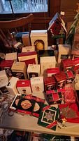 Wide photo of multiple Hallmark Keepsake ornaments in boxes and loose on a table with chairs and window in background.