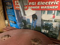 Power washer box showing 1300 PSI label and brand I-CELL with visible wear and tear, also showing corner of round wood table beneath.