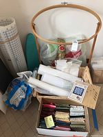 Photo shows large wooden quilting hoop, rolls of fabric and batting, basket, and various quilting supplies including small packaged materials.