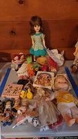 Full lot overview showing multiple dolls including Raggedy Ann and Andy, trolls, large vintage doll in blue floral dress, small dolls, and plastic toys on table