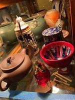 Photo showing multiple items: green pottery vase, ceramic stein with lid, pottery jam jar with lid and spoon, red and blue ceramic bowl, brown ceramic teapot, decorative glass item with brass spoon.