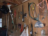 Wide view of pegboard holding string trimmer, hand tools including hammers, saws, crowbar, and bundled metal wires plus a plastic bag containing additional items