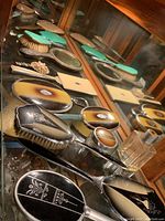 Glass cabinet shelf showing assorted vintage vanity pieces including ombre brown celluloid mirrors and brushes, mint green enameled brush set, silver plated brush, black and white enamel tray and clear glass container.