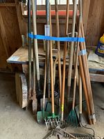 Photo showing multiple yard tools leaning against a shelf, including shovels, hole diggers, spading forks, and rakes. Tools have wooden handles and metal heads with visible rust and dirt.