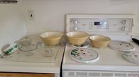 Overview of the three cream vintage mixing bowls and accompanying glass dishes including pedestal, baking, and dessert dishes placed on kitchen counter and stove.
