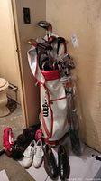 Full view of golf set in Wilson golf bag with multiple clubs and men's white golf shoes and brown dress shoes at base