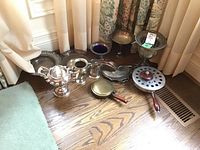 Overview of silver plate kitchenware including trays, bowls, buckets, and bed warmer on wooden floor near curtain.