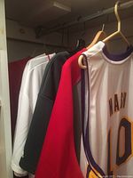 View of five sports jerseys and shirts hanging in a closet, showing the side of a Steve Nash Lakers basketball jersey and other shirts behind it.