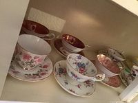 Overview of seven assorted vintage teacups and saucers on shelf showing various floral patterns and gold trims.