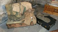Two vintage military trunks with various beige and khaki military clothing items including jackets and pants draped over and inside them