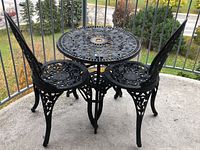 View of the black metal bistro set showing the round table and two chairs with ornate openwork design, placed on a concrete balcony floor.