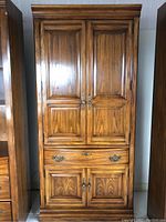 Tall oak cabinet with two doors, polished finish, brass handles, and raised panel design.