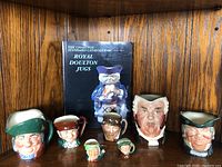 Full view of six Toby and character jugs arranged on a wooden shelf with The Charlton Standard Catalogue of Royal Doulton Jugs behind them, showing the variety of shapes, sizes, and colors in the lot.