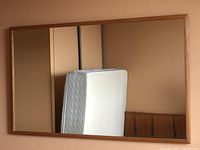 Full front view of large rectangular mirror reflecting a mattress and wall panel. Frame is teak wood with natural medium brown finish.