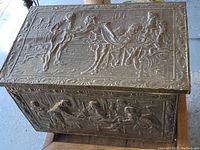 Top view showing embossed brass panel with a detailed old world scene of four people in period attire interacting indoors.