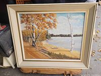 Frontal view of the framed shoreline painting showing autumn trees by water with sandy shore, partly cloudy sky, and a wooden frame.