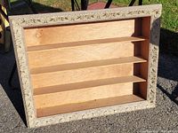 Front perspective of the knick knack shelf made from an old ornate picture frame with three shelves inside, placed outdoors.