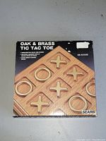 Front view of sealed product box showing tic tac toe board with oak base and brass pieces, labeled 'Oak & Brass Tic Tac Toe' from Sears.