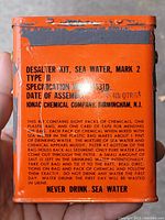 Close up photo of the front side of an orange metal tin container labeled Desalter Kit, Sea Water, Mark 2, Type II with chemical instructions and warning text.