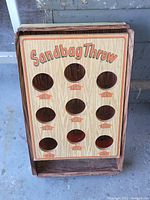 Front-facing photo showing the tin sandbag throw game inside a wooden frame, clearly displaying nine point-labeled holes for throwing sandbags.