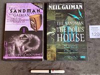 Front covers of two graphic novels - 'Preludes & Nocturnes' and 'The Doll's House' by Neil Gaiman's The Sandman series, placed side by side with a branded pen for scale.