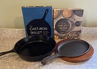 Two black cast iron skillets with two cookbooks standing behind them, one skillet on wooden hot plate