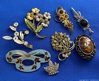 Eight assorted vintage brooches displayed on blue fabric, showing floral designs, enamel, rhinestones, and metal work.