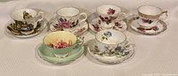 View of all seven fine china tea cup and saucer sets arranged with different floral and cottage patterns.