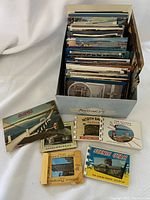 Box filled with assorted vintage postcards standing upright showing a variety of colorful card backs and fronts, some displayed on the table around the box