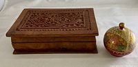 Rectangular wood carved keepsake box next to small round ornate ring box on white surface