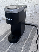 Front diagonal view of black Keurig K-Mini coffee maker machine, plugged in, sitting on a striped tablecloth against white textured wall.