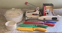 Photo showing large Corning Ware casserole dish with lid, a small ramekin, four color-coded Cuisinart knives with protective sheaths, white KitchenAid blending stick, manual french fry cutter, and a boxed rotary cheese grater.