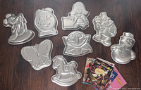 Eight vintage Wilton cake pans arranged on dark wood floor including Elmo, The Count, Yosemite Sam, Mickey Mouse, butterfly, rocking horse, snowman, and clock along with four vintage cake decorating books.
