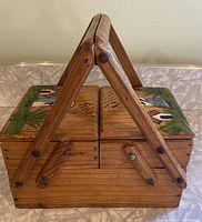 View of closed wooden picnic basket with hand painted scene on lid depicting houses, a woman, and greenery. Basket has two handles and wood construction.
