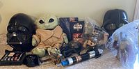 Photo showing full-size Darth Vader helmet, smaller Darth Vader helmet figure, plush Grogu doll, Star Wars action figures, toy light saber, packaged items including trading cards and a Funko Pop figure.