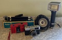 Photo shows two vintage cameras, a tripod in its Vivitar box, and a black and silver flash with accessory flash bulb and cord