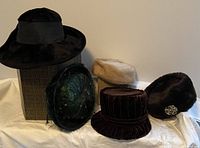 Five vintage ladies hats displayed together with a hat box, showing variety in textures and decorations including velvet, faux fur, feather, and brooch details.