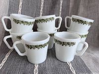 Eight white milk glass coffee mugs with green floral band around the rim arranged in groups.