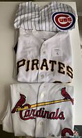 Three baseball jerseys stacked on a white surface showing Cubs, Pirates, and Cardinals logos clearly.