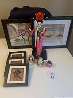 Photo showing entire lot: Asian doll, framed art prints, smaller framed calligraphy prints, and figurines on a white surface